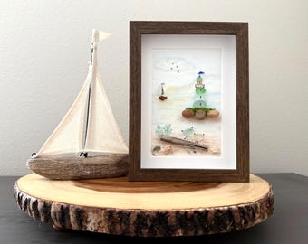 May include: A framed artwork depicts a lighthouse and two figures on a beach, crafted from pebbles. A wooden sailboat with a white sail and a wooden base sits to the left. The artwork and sailboat are displayed on a wooden slice.