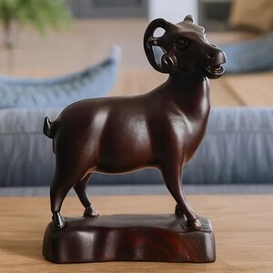 May include: A dark brown wooden ram sculpture. The ram is standing on a rectangular base, with detailed horns and a smooth, polished finish. The sculpture is placed on a wooden table in a living room setting.