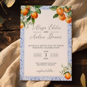 Puede incluir: Invitación de boda con ilustraciones de acuarela de naranjas y flores. La invitación tiene un borde decorativo azul y blanco e incluye los nombres Alussa Edelen y Andrew Donner, junto con los detalles de la boda.