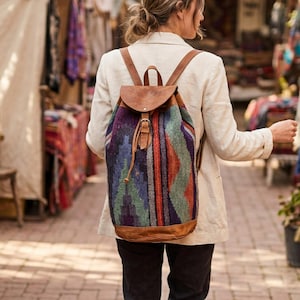 May include: A colorful backpack with a geometric pattern in shades of purple, blue, orange, and green. The backpack has brown leather straps, a flap closure, and a base. The person is walking in a market.