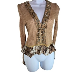 Peut inclure: Cardigan beige &agrave; manches longues et col en V. Le col et le panneau central sont orn&eacute;s de perles et de broderies. Le bas pr&eacute;sente un ourlet &agrave; volants avec un tissu &agrave; motifs. Le cardigan se ferme avec des boutons.