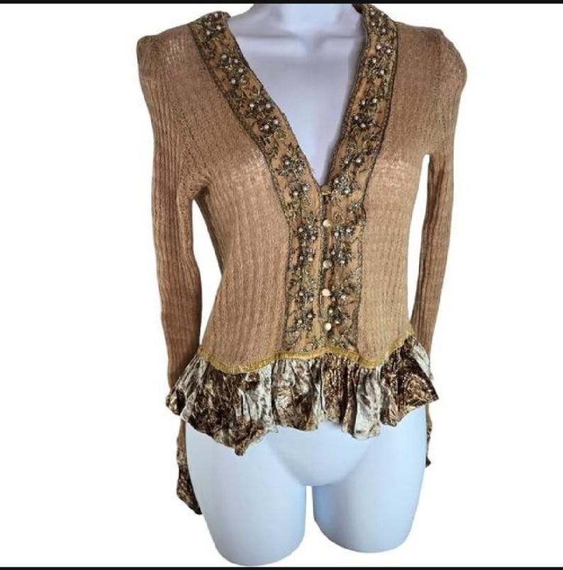 Peut inclure: Cardigan beige &agrave; manches longues et col en V. Le col et le panneau central sont orn&eacute;s de perles et de broderies. Le bas pr&eacute;sente un ourlet &agrave; volants avec un tissu &agrave; motifs. Le cardigan se ferme avec des boutons.