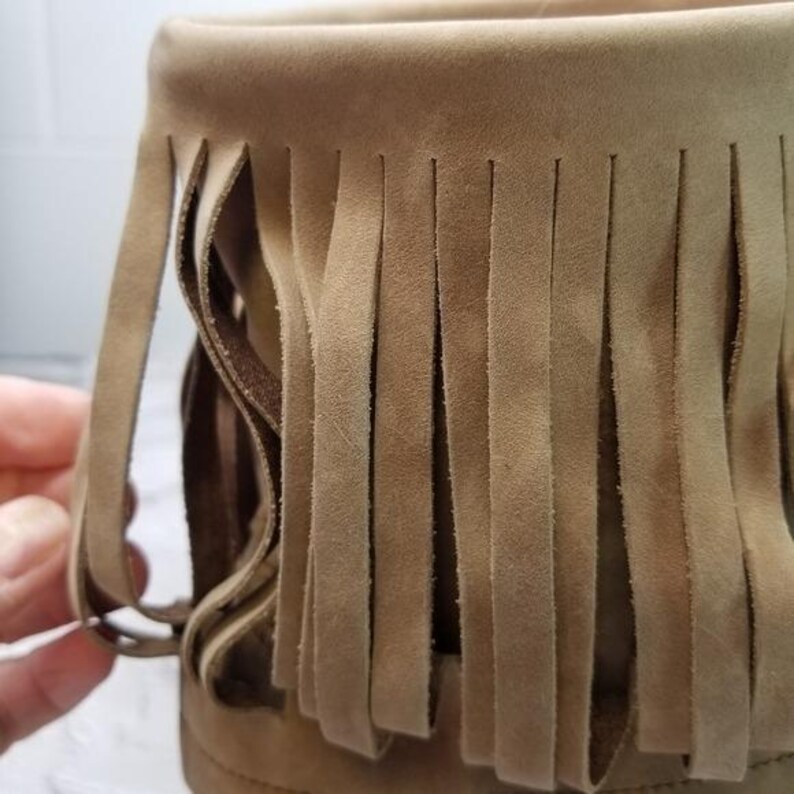 May include: Close-up of a tan suede boot with a fringed design. The fringe hangs vertically from the top of the boot, creating a textured and stylish look. The boot appears to be made of soft suede material.