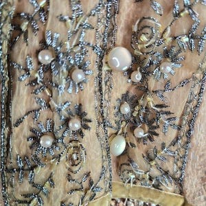 Peut inclure: Gros plan sur un v&ecirc;tement p&ecirc;che avec des broderies argent&eacute;es complexes et des perles. Le motif pr&eacute;sente des motifs floraux et de feuilles, avec de gros boutons ronds en forme de perles. Le tissu est d&eacute;licat et transparent.