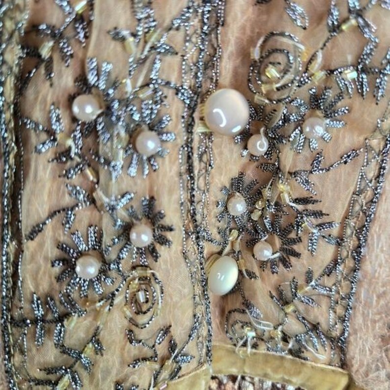 Peut inclure: Gros plan sur un v&ecirc;tement p&ecirc;che avec des broderies argent&eacute;es complexes et des perles. Le motif pr&eacute;sente des motifs floraux et de feuilles, avec de gros boutons ronds en forme de perles. Le tissu est d&eacute;licat et transparent.