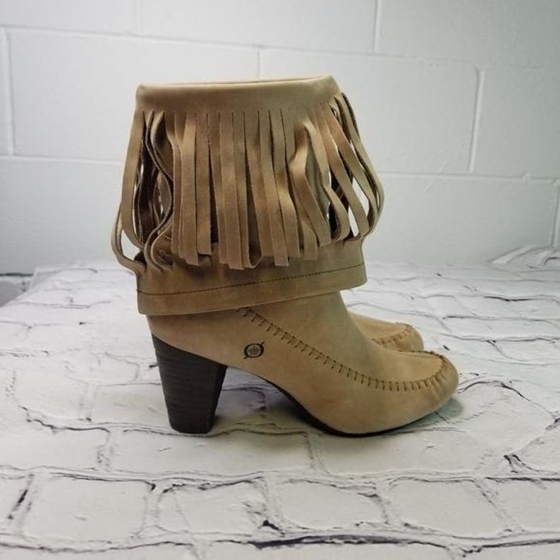 May include: Tan suede ankle boots with a fringe detail around the top. The boots have a rounded toe, a stacked heel, and decorative stitching. The boots are approximately 3 inches in heel height.