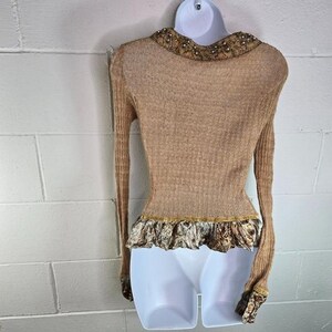 Peut inclure: Pull &agrave; manches longues de couleur beige avec un col d&eacute;coratif et un ourlet &agrave; volants. Le col est orn&eacute; de petits accents scintillants. L'ourlet pr&eacute;sente un tissu &agrave; motifs avec une bordure dor&eacute;e. Le pull est sur un mannequin blanc.