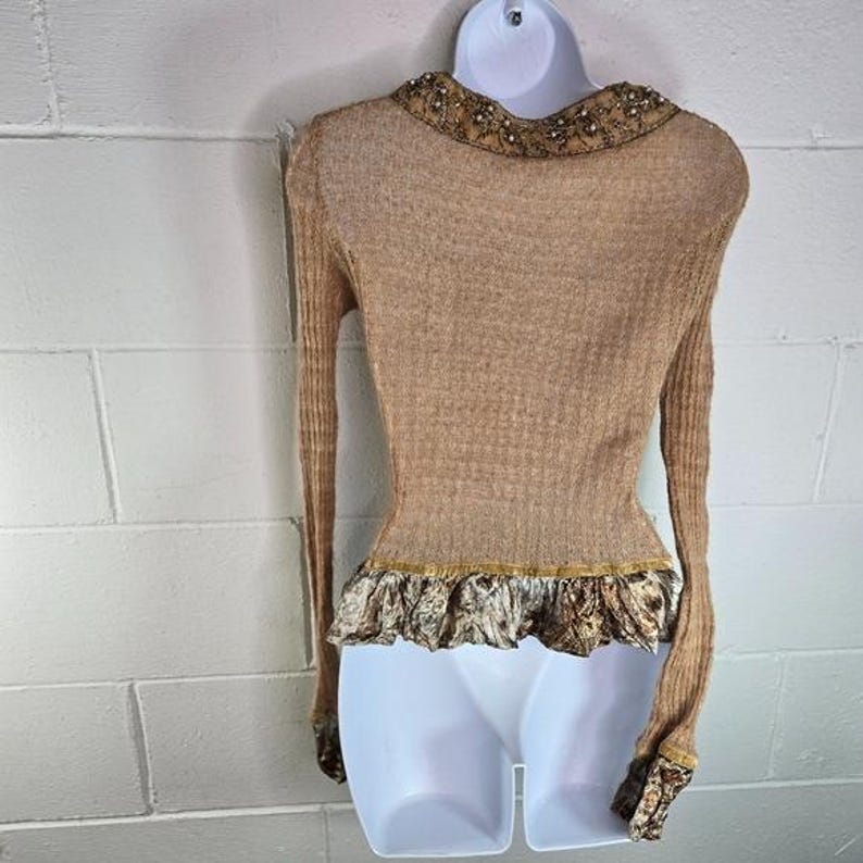 Peut inclure: Pull &agrave; manches longues de couleur beige avec un col d&eacute;coratif et un ourlet &agrave; volants. Le col est orn&eacute; de petits accents scintillants. L'ourlet pr&eacute;sente un tissu &agrave; motifs avec une bordure dor&eacute;e. Le pull est sur un mannequin blanc.
