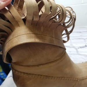 May include: Close-up of a tan leather boot with a decorative fringe around the ankle. The boot has a low heel and visible stitching details. The leather appears worn, giving it a vintage look.