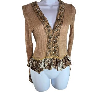 Peut inclure: Cardigan beige &agrave; col en V et manches longues. L'encolure et le panneau central sont orn&eacute;s de perles et de broderies. Le bas pr&eacute;sente un ourlet &agrave; volants avec un tissu &agrave; motifs.