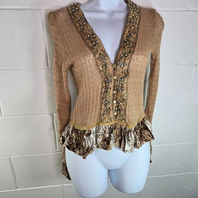 Peut inclure: Un cardigan beige &agrave; manches longues et col en V. L'encolure et le panneau central sont orn&eacute;s de perles et de sequins. Le bas pr&eacute;sente un ourlet &agrave; volants avec un tissu &agrave; motifs. Le cardigan est pr&eacute;sent&eacute; sur un mannequin blanc.