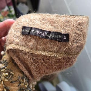 Peut inclure: Gros plan sur un tissu duveteux de couleur beige avec une &eacute;tiquette noire portant l'inscription "hazel". Le tissu semble faire partie d'un v&ecirc;tement, peut-&ecirc;tre une manche ou un poignet, avec des perles complexes visibles sur le bord.