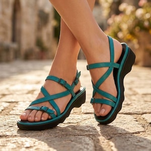 May include: Teal leather sandals with crisscross straps and adjustable buckles. The sandals have a black sole and a slight wedge heel. The image shows the sandals on a cobblestone surface.