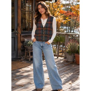 May include: A sleeveless button-front vest featuring a dark blue and rust orange plaid pattern, worn over a long-sleeved cream-colored henley shirt with a button placket, paired with light blue wide-leg denim jeans.