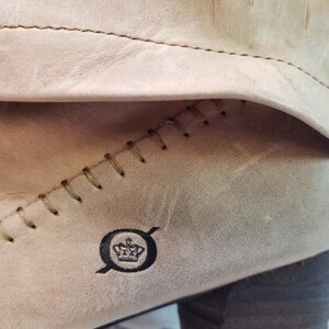 May include: Close-up of a light tan leather boot. Visible are the stitched seams, the brand logo, and the texture of the leather. The stitching is a contrasting olive green color.