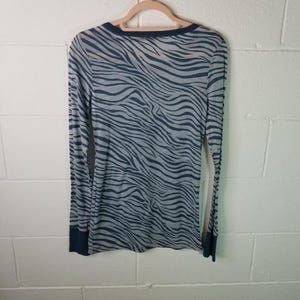 May include: A long-sleeved top with a zebra stripe design in grey and dark blue. The garment is suspended on a wooden hanger against a white brick wall. The cuffs and neckline are dark blue.