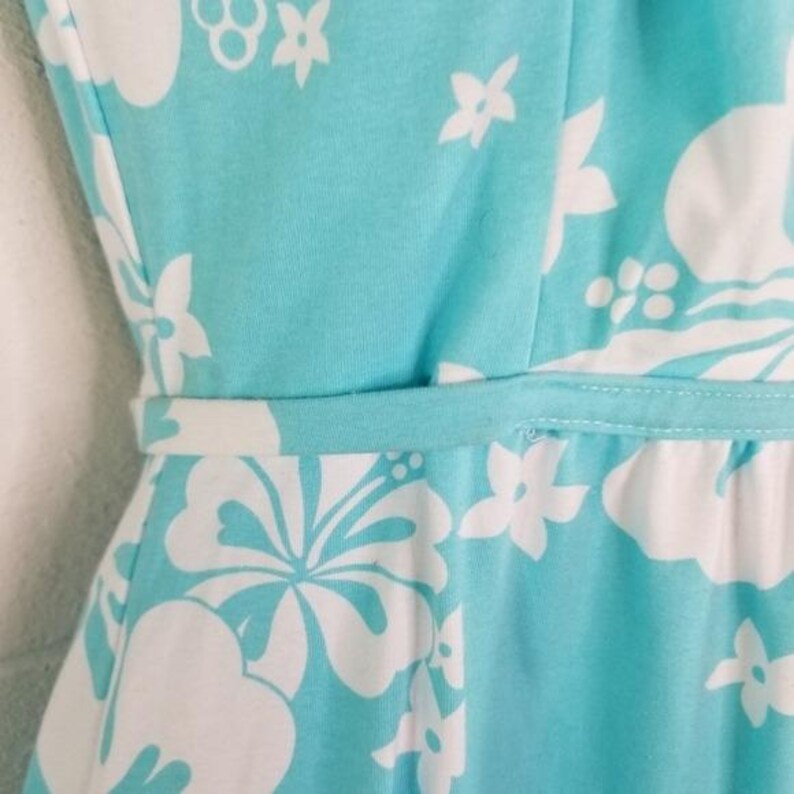 May include: A sleeveless turquoise dress with a white floral pattern. The dress features a fitted bodice and a waist belt. The fabric is a soft, lightweight material, perfect for warm weather. The floral design includes hibiscus and other tropical flowers.
