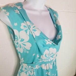 May include: A sleeveless turquoise dress with a white floral pattern. The dress features a cowl neckline and a fitted waist. The fabric is lightweight and appears to be a soft material. The dress is displayed on a white mannequin.