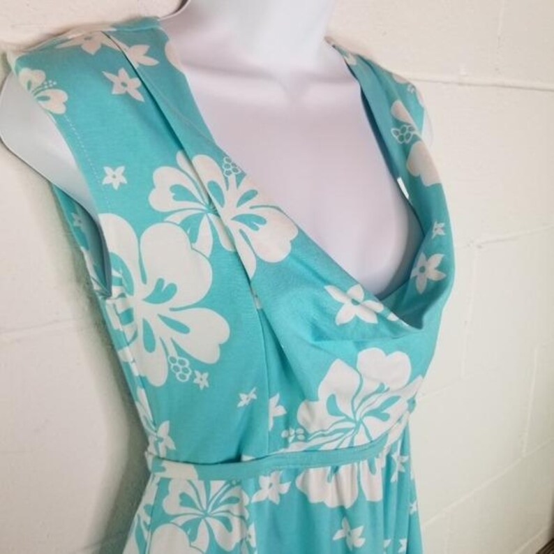 May include: A sleeveless turquoise dress with a white floral pattern. The dress features a cowl neckline and a fitted waist. The fabric is lightweight and appears to be a soft material. The dress is displayed on a white mannequin.