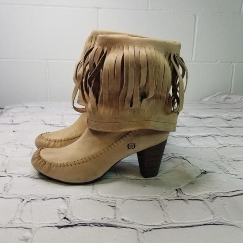 May include: A pair of tan suede ankle boots with fringe detailing. The boots have a rounded toe, a stacked heel, and a stitched design along the seams. The boots are approximately 3 inches (7.6 cm) high.