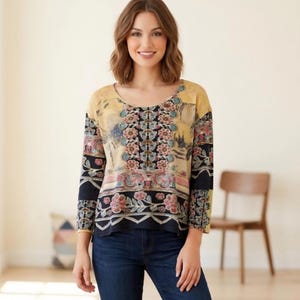 May include: A long-sleeved top with a scoop neck, featuring a floral pattern in shades of yellow, black, pink, and blue. The top is paired with blue jeans. The design includes a detailed floral border at the hem and sleeves.