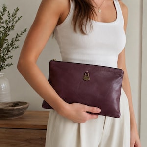 May include: A deep burgundy leather clutch with a brass-colored clasp and zipper. The rectangular bag is held by a person wearing a white tank top and cream-colored pants. The clutch has a smooth, slightly textured surface.