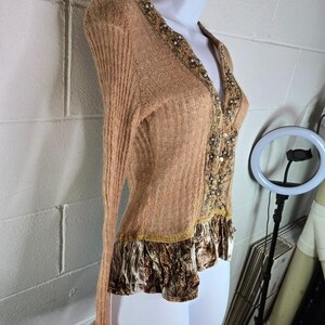Peut inclure: Cardigan beige &agrave; manches longues et col en V. Le devant et l'encolure sont orn&eacute;s de perles et d'un tissu &agrave; motifs. L'ourlet inf&eacute;rieur pr&eacute;sente une bordure &agrave; volants avec un motif marron et cr&egrave;me. Le cardigan est pr&eacute;sent&eacute; sur un mannequin.