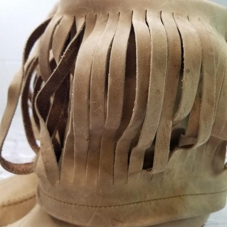 May include: Close-up of a tan suede boot with a fringed top. The fringe is made of long, thin strips of the same material. The boot has a stitched seam detail.