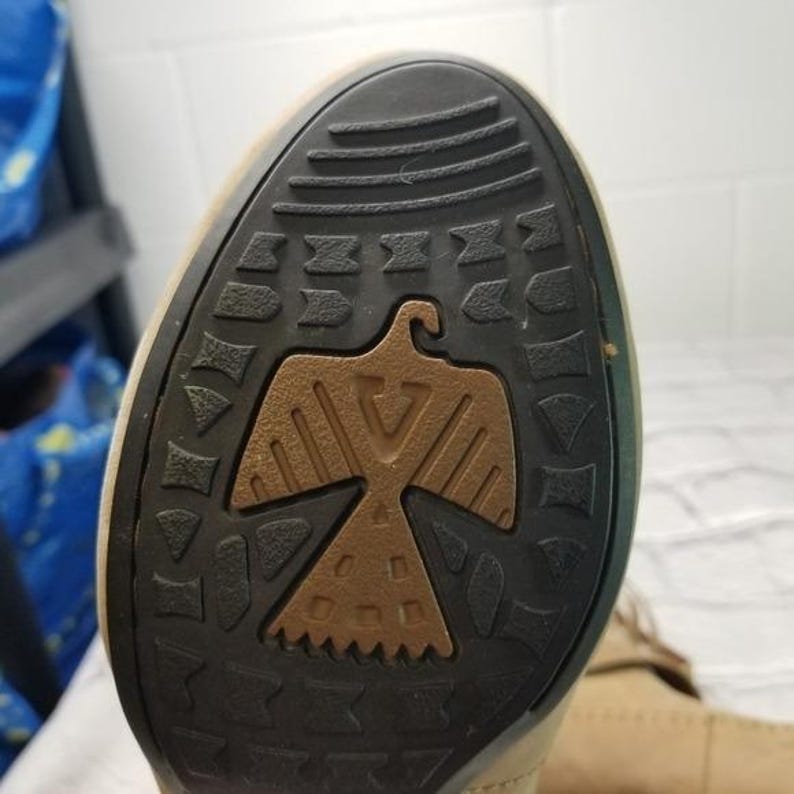 May include: Close-up of the sole of a tan boot. The sole is black with a brown eagle design in the center. The eagle is stylized with outstretched wings. The boot's sole has a textured pattern for grip.