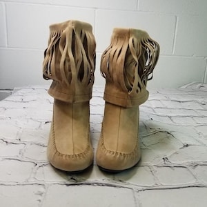 May include: A pair of tan suede boots with fringe detailing. The boots have a rounded toe and a stacked heel. The fringe is around the top of the boot. The boots are sitting on a white brick surface.