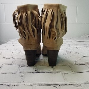May include: A pair of tan suede ankle boots with a fringe design. The boots have a stacked heel and are displayed against a white brick wall background.