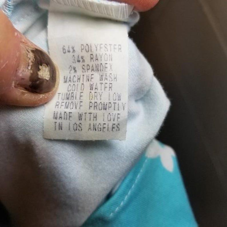 May include: A white clothing tag with black text, held by a hand. The tag reads: 64% POLYESTER, 34% RAYON, 2% SPANDEX, MACHINE WASH COLD WATER, TUMBLE DRY LOW, REMOVE PROMPTLY, MADE WITH LOVE IN LOS ANGELES.