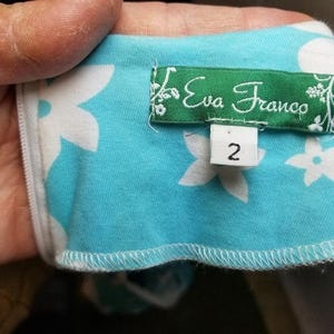 May include: A close-up of a piece of fabric with a floral pattern in white on a light blue background. A green label with the text "Eva Franco" and a size 2 tag are visible.