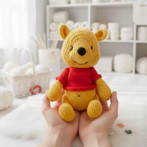Peut inclure: Une peluche Winnie l'ourson faite à la main au crochet. L'ours est jaune avec un haut rouge et un nez noir. Le jouet est tenu dans les mains, avec une surface blanche et moelleuse en arrière-plan. L'ours a une expression souriante.