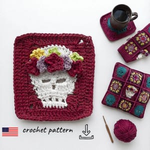 May include: A square crochet motif featuring a white skull adorned with a colourful floral crown in shades of yellow, green, and purple, set against a deep burgundy background. Nearby are a matching coaster, a small square pouch, and yarn.