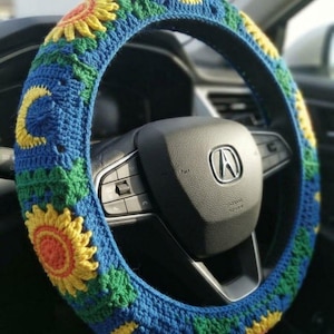 Puede incluir: Una funda de volante de ganchillo con un patrón repetitivo de girasoles amarillos con centros naranjas y lunas crecientes amarillas sobre un fondo azul con detalles de hojas verdes, colocada sobre un volante negro con un logo Acura plateado.