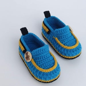 Può includere: Un paio di scarpine da neonato all'uncinetto blu fatte a mano con dettagli gialli e neri. Ogni scarpa presenta un bottone in legno con un disegno di orsetto e una linguetta nera. Le scarpe misurano circa 10 cm.