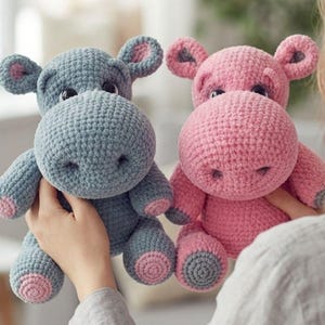 May include: Two crocheted hippopotamus stuffed animals. One hippo is grey with pink accents on the ears and feet, while the other is pink with grey accents on the ears and feet. Both have dark, round eyes and a textured, yarn-based surface.