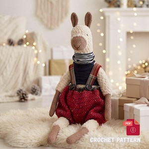 May include: A handmade crochet llama doll with a cream-colored body, brown ears, and a brown muzzle. It wears a navy blue sweater with a striped collar and red overalls. The doll is sitting on a white rug.