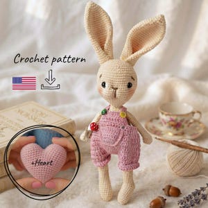 Puede incluir: Un muñeco de conejo de ganchillo beige con un peto rosa, botones pequeños y un accesorio de seta roja. La imagen incluye el texto Crochet pattern, un icono de descarga, una bandera de EE. UU. y un recuadro con un corazón rosa y +Heart.
