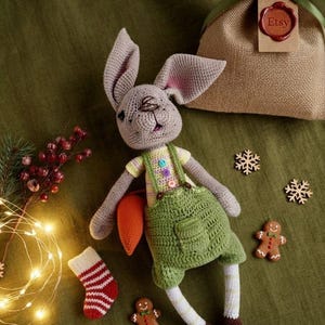Peut inclure: Un lapin en crochet fait de fil gris, portant un t-shirt rayé jaune avec trois boutons colorés et une salopette verte. Le lapin tient une carotte orange et possède des jambes rayées blanc et jaune avec des pieds marron, réalisé en détail.