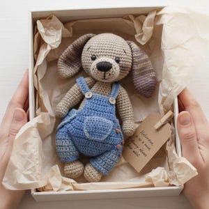 May include: A handmade crochet dog toy in a white box. The dog is beige with long ears and a black nose, wearing blue dungarees. The box is lined with brown paper. A small tag is attached with a wooden peg.