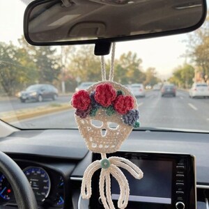 May include: A white crocheted skull ornament hanging from a rearview mirror. The skull features a crown of pink, purple, and teal crocheted flowers across the forehead and a light blue button at the base with dangling white crocheted ribbons.