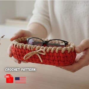 May include: A red and tan crochet basket with a bow, holding a pair of black-framed glasses. The basket is oval-shaped and has a tan trim. The text "CROCHET PATTERN" is visible.