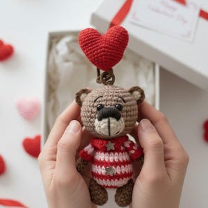 May include: A crocheted teddy bear with a red heart above its head. The bear has a brown body, a red and white striped shirt, and brown paws. The bear is held in two hands, with a gift box in the background.