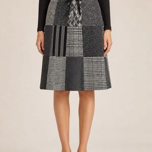 May include: A knee-length patchwork skirt in shades of gray, black, and white. The skirt features a variety of textured fabrics, including plaid, herringbone, and solid panels. A black drawstring detail is at the waist. The person is wearing black loafers and a black long-sleeve shirt.