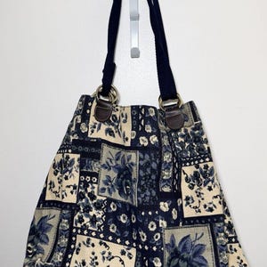 May include: A tote bag featuring a patchwork design of blue and cream floral patterns. The bag has two dark blue fabric shoulder straps attached with gold-toned metal rings and brown leather-like tabs at the top edge of the bag.