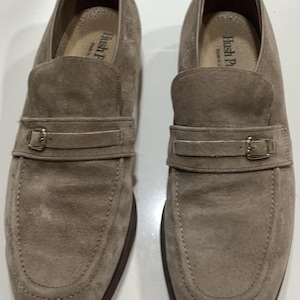 May include: A pair of light brown suede loafers with a buckle detail. The shoes have a rounded toe and a dark brown sole. The brand name "Hush Puppies" is visible on the insole.