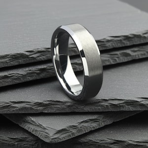 May include: A silver ring with a brushed metal finish. The ring features a flat top and beveled edges, offering a modern, minimalist design. The ring is presented on a dark grey, textured surface.