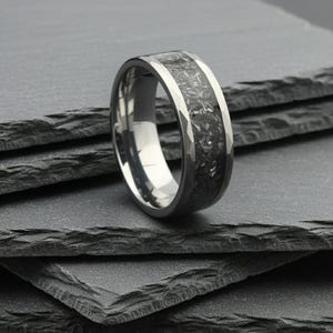 Puede incluir: Un anillo plateado con una banda central oscura y texturizada. El anillo tiene un borde exterior facetado y un interior pulido, mostrando un diseño moderno. Esta joya es adecuada para el uso diario.